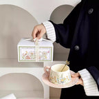 Person holding a floral teacup and saucer with a matching box on a neutral background
