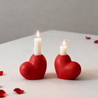 Two red heart-shaped candle holders with lit candles on a light surface with scattered red petals.
