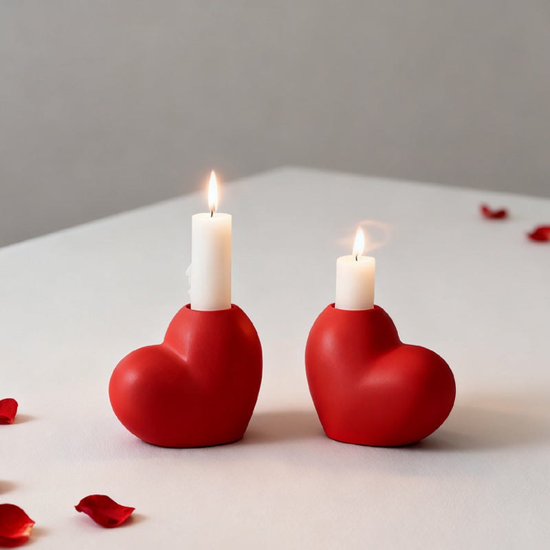 Two red heart-shaped candle holders with lit candles on a light surface with scattered red petals.
