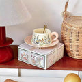 Decorative teacup and saucer set on a box with a wicker basket and red lamp in the background.