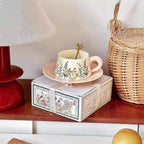 Decorative teacup and saucer set on a box with a wicker basket and red lamp in the background.